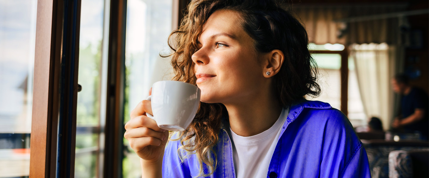 Junge Frau sitzt im Café mit Tasse in der Hand und schaut verträumt aus dem Fenster.