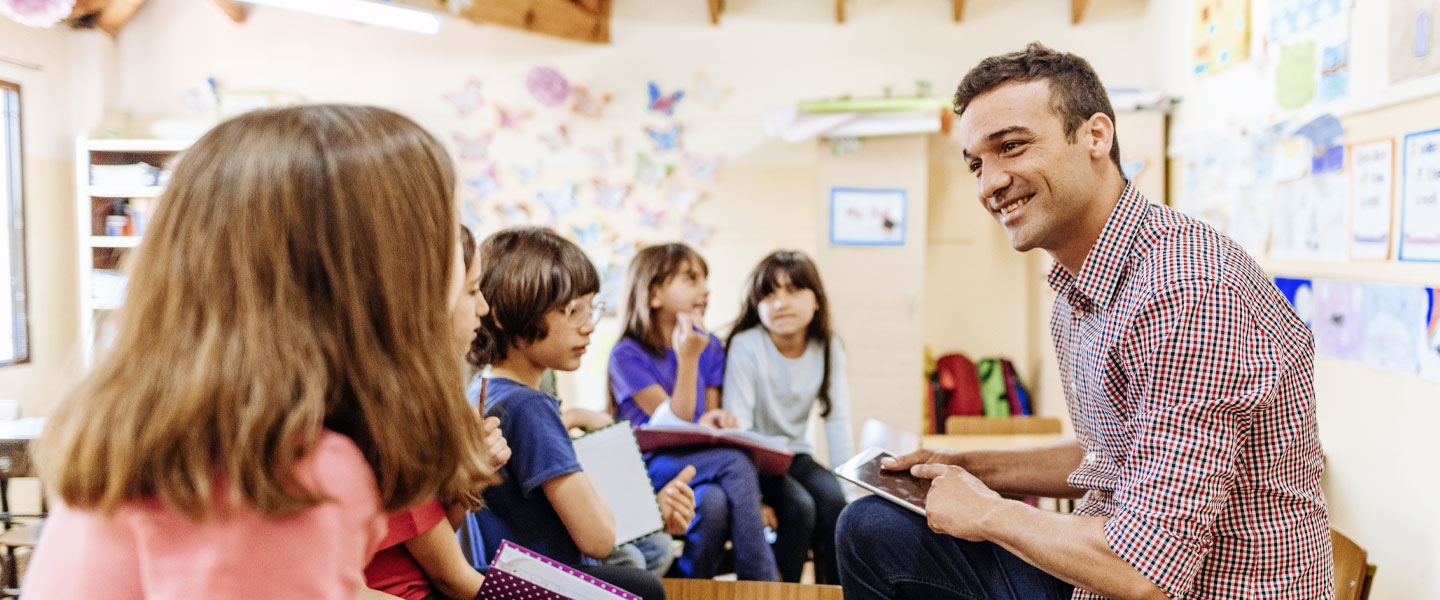 Ein Lehrer sitzt vor einer Gruppe an Kindern.