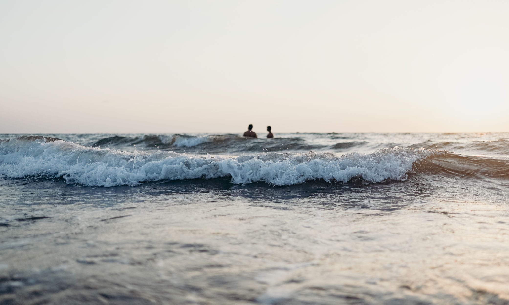 zwei Badende im Meer, fotografiert vom Strand vor anrauschenden Brandung und bei Sonnenuntergang. Wie man sich diesen Traum mit einer Reiserücktrittversicherung auch gegen Corona bewahren kann